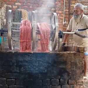 May include: A man operates a machine dyeing fabric. The machine has large metal wheels and is set over a brick structure. Red fabric is draped over the wheels, with steam rising. The background features a brick wall and other dyed fabrics.
