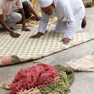 May include: A rectangular woven rug featuring a light green and white checkered pattern with pink floral accents at the corners. In the foreground, piles of loose, unspun yarn in shades of red, olive green, and cream are arranged on the ground.