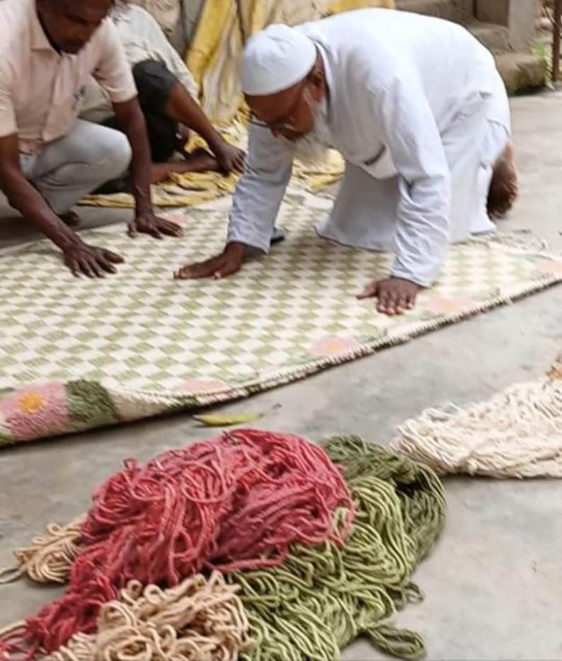 May include: A rectangular woven rug featuring a light green and white checkered pattern with pink floral accents at the corners. In the foreground, piles of loose, unspun yarn in shades of red, olive green, and cream are arranged on the ground.