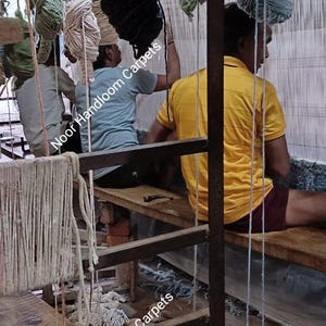 May include: A traditional handloom setup with multiple skeins of yarn in shades of peach, sage green, cream, black, and blue hanging from a wooden frame. The text Noor Handloom Carpets is visible across the image, which shows the weaving process.