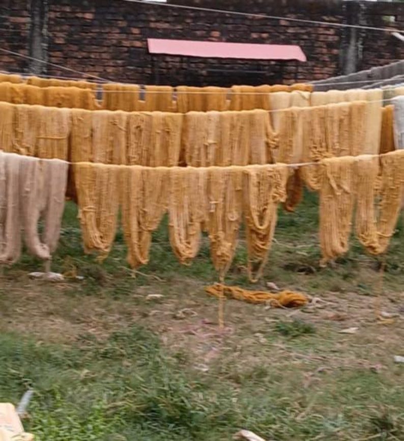 May include: Multiple long, thin hanks of golden-yellow and light beige yarn are draped over horizontal white lines to dry. The strands of fiber hang vertically in rows, creating a repetitive pattern of textured, fibrous material outdoors.