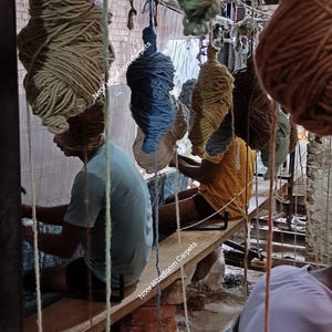 May include: Image of a carpet weaving workshop. Several large bundles of yarn in various colors hang from the ceiling. Workers are seated at wooden looms, actively weaving. The text "Noor Handloom Carpets" is visible.