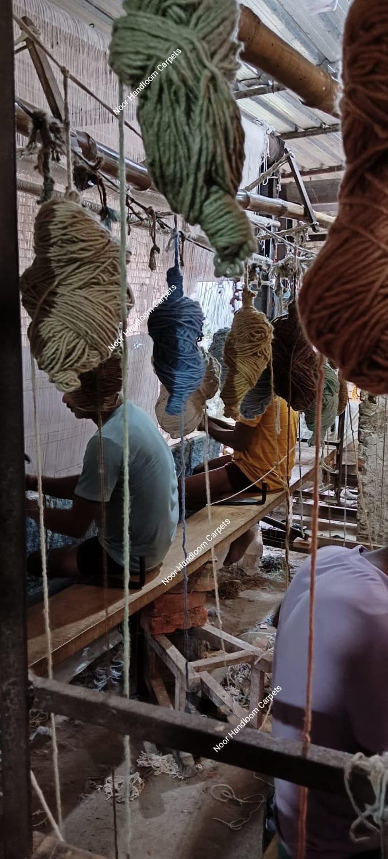 May include: Image of a carpet weaving workshop. Several large bundles of yarn in various colors hang from the ceiling. Workers are seated at wooden looms, actively weaving. The text "Noor Handloom Carpets" is visible.