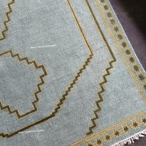May include: A light blue area rug featuring a dark green and yellow geometric zigzag border pattern. The rug has a textured surface with a repeating star-like motif along the edge and white fringe at the bottom. The text Noor Handloom Carpets is visible.