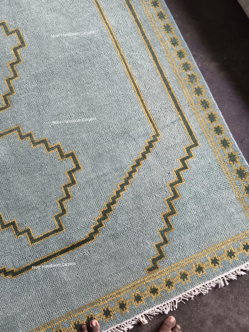 May include: A light blue area rug featuring a dark green and yellow geometric zigzag border pattern. The rug has a textured surface with a repeating star-like motif along the edge and white fringe at the bottom. The text Noor Handloom Carpets is visible.