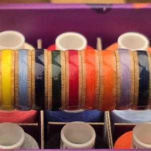 Puede incluir: Una variedad de carretes de hilo coloridos, cada uno con un color de hilo diferente. Los carretes están dispuestos en una caja con compartimentos individuales, y el hilo está enrollado alrededor de carretes de madera.