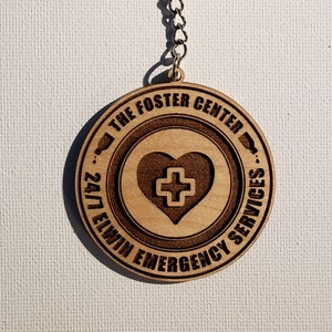 May include: Wooden keychain with a brown heart and a white cross inside. The keychain has the text "The Foster Center 24/7 Elwin Emergency Services" around the heart.
