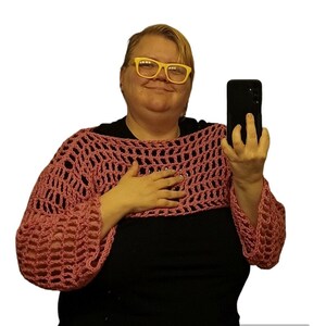 May include: A pink crocheted top with long sleeves. The top has a loose, open weave and is worn over a black shirt.
