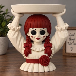 Può includere: Una statuetta color crema di una bambola con capelli rossi intrecciati, che indossa un vestito con volant e una rosa rossa. La bambola ha grandi occhi neri e tiene un vassoio circolare sopra la testa con entrambe le mani come espositore.