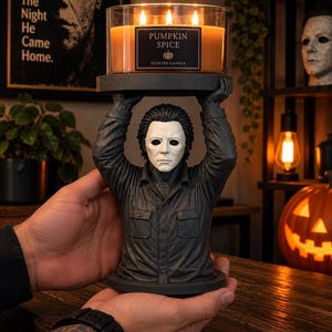 Könnte beinhalten: Eine dreidochtige Pumpkin Spice Duftkerze in einem klaren Glasgefäß, platziert auf einem dunkelgrauen Kerzenhalter in Form einer Büste mit weißer Maske und dunklem Haar, bekleidet mit einem dunklen Hemd, die Arme erhoben, um den Kerzensockel zu stützen.