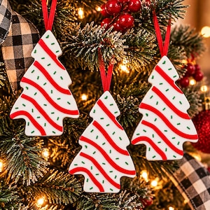 May include: Three white Christmas tree ornaments featuring diagonal red wavy stripes and small green sprinkles. Each ornament is shaped like a stylized evergreen tree and hangs from a thin red ribbon loop against a pine tree background.