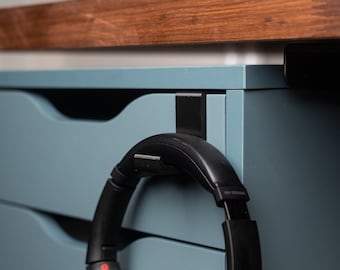 May include: Black over-ear headphones with a padded headband and earcups, hanging on a black hook attached to a light blue cabinet.