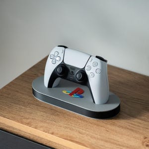 May include: A white PlayStation 5 controller is sitting on a grey and black charging stand with the PlayStation logo on the front. The stand is on a wooden surface.