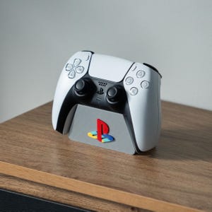 May include: A white PlayStation 5 controller is sitting on a grey stand with the PlayStation logo in red, yellow and blue. The stand is on a wooden surface.