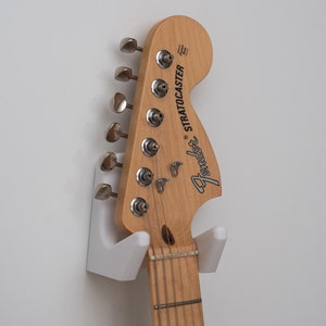 May include: A white guitar wall mount with a brown Fender Stratocaster guitar hanging on it.