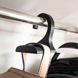 May include: A black plastic hanger with a hook designed to hold a purse or handbag. The hanger is hanging from a metal rod and has a brown leather strap attached to it.