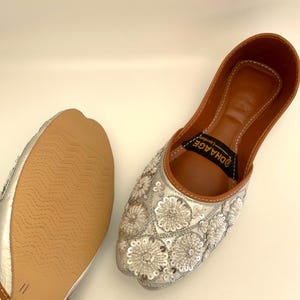 Peut inclure: Une paire de chaussures plates argent&eacute;es brod&eacute;es avec une semelle int&eacute;rieure en cuir marron. Les chaussures ont un motif floral avec des paillettes et le texte "DHAAGE" est imprim&eacute; sur la semelle int&eacute;rieure.