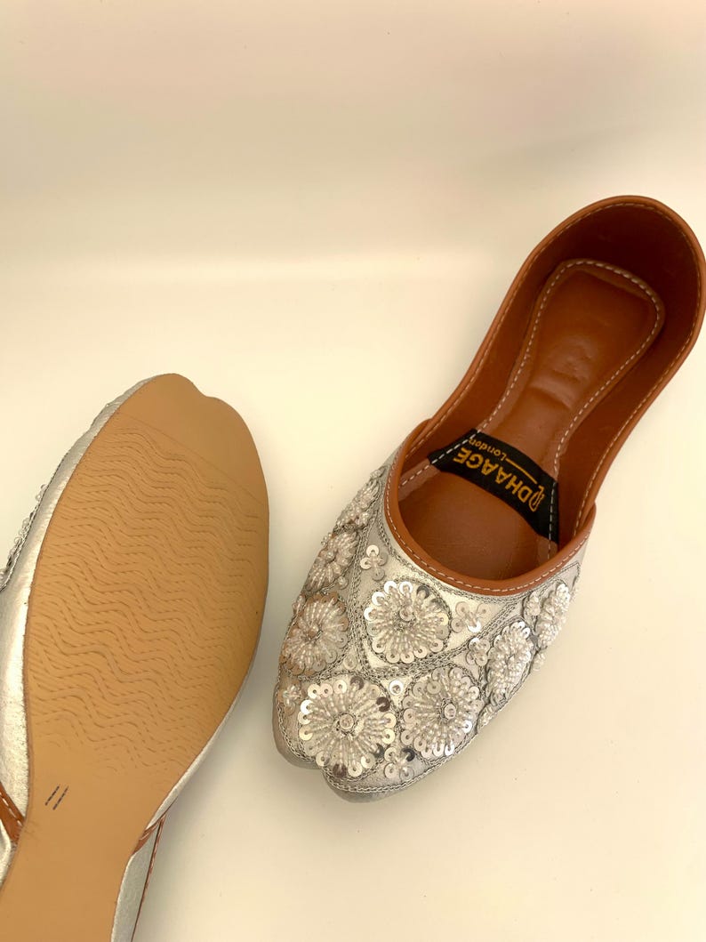 Peut inclure: Une paire de chaussures plates argent&eacute;es brod&eacute;es avec une semelle int&eacute;rieure en cuir marron. Les chaussures ont un motif floral avec des paillettes et le texte "DHAAGE" est imprim&eacute; sur la semelle int&eacute;rieure.