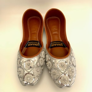 Peut inclure: Une paire de chaussures plates brod&eacute;es argent&eacute;es avec une doublure en cuir marron. Les chaussures ont un motif floral avec des paillettes et le nom de la marque "DHAAGE London" est imprim&eacute; &agrave; l'int&eacute;rieur.