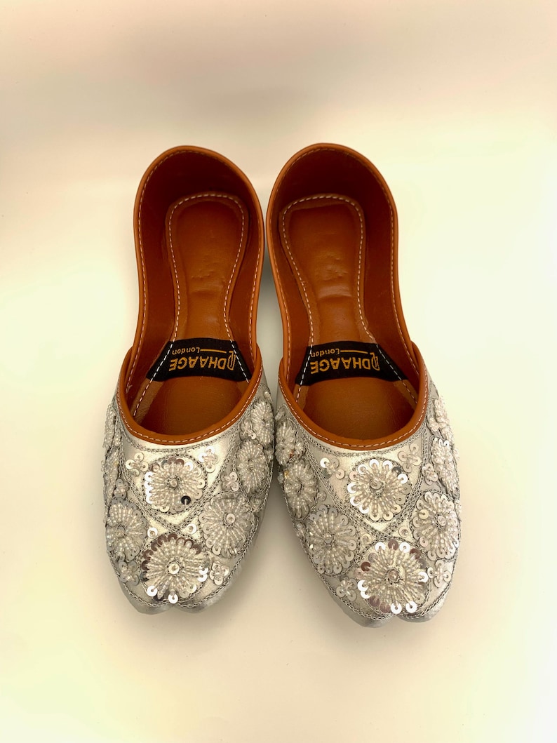 Peut inclure: Une paire de chaussures plates brod&eacute;es argent&eacute;es avec une doublure en cuir marron. Les chaussures ont un motif floral avec des paillettes et le nom de la marque "DHAAGE London" est imprim&eacute; &agrave; l'int&eacute;rieur.