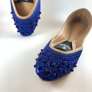 May include: A pair of blue and gold embellished flat shoes with a beaded design. The shoes have a pointed toe and a low heel. The brand name "D'HAGE" is visible on the shoes.