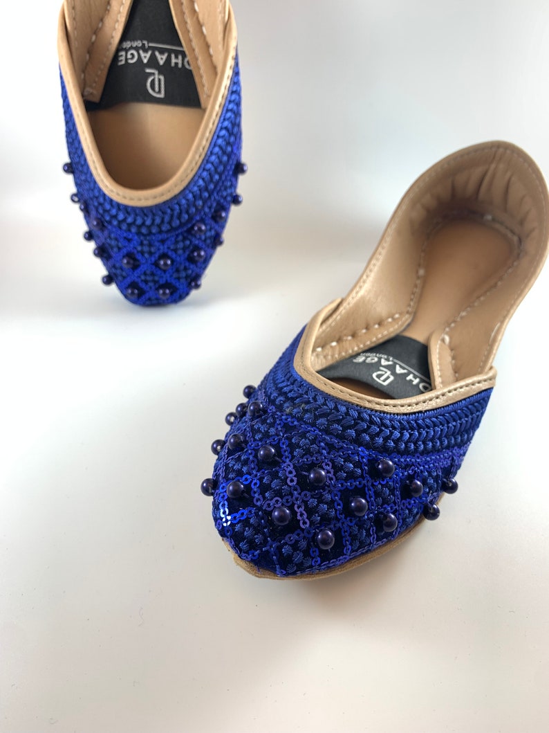 May include: A pair of blue and gold embellished flat shoes with a beaded design. The shoes have a pointed toe and a low heel. The brand name "D'HAGE" is visible on the shoes.