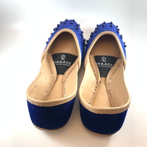 May include: A pair of blue velvet shoes with gold trim and beaded embellishments. The shoes have a pointed toe and a low heel. The brand name "DHAAGE London" is printed on the insole.