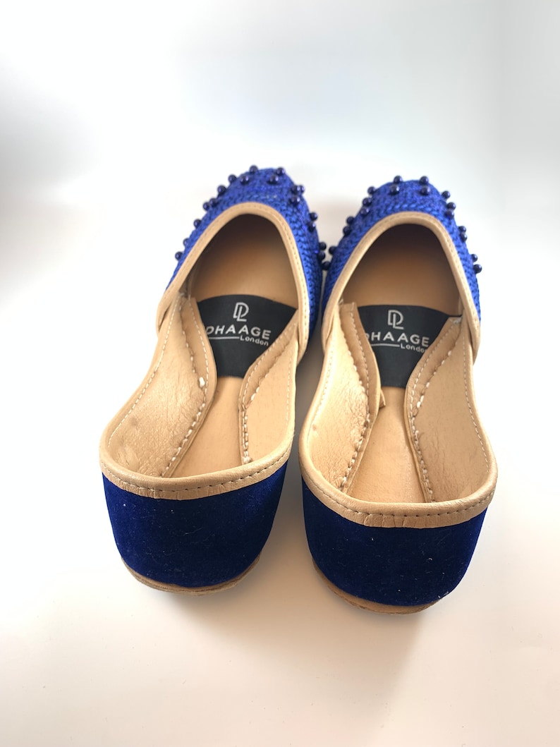 May include: A pair of blue velvet shoes with gold trim and beaded embellishments. The shoes have a pointed toe and a low heel. The brand name "DHAAGE London" is printed on the insole.