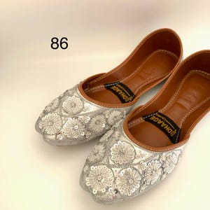 Peut inclure: Une paire de chaussures plates argent&eacute;es brod&eacute;es avec une doublure en cuir marron. Les chaussures ont un motif floral avec des paillettes et le nom de la marque "DHAAGE" est imprim&eacute; sur la doublure.