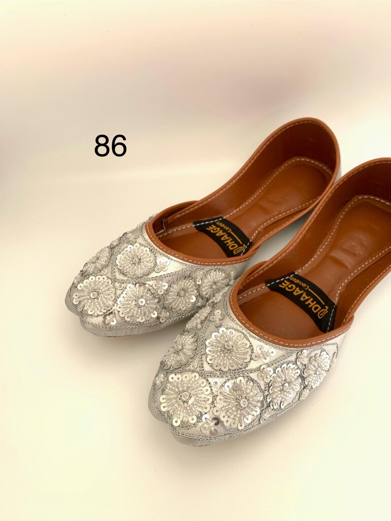 Peut inclure: Une paire de chaussures plates argent&eacute;es brod&eacute;es avec une doublure en cuir marron. Les chaussures ont un motif floral avec des paillettes et le nom de la marque "DHAAGE" est imprim&eacute; sur la doublure.