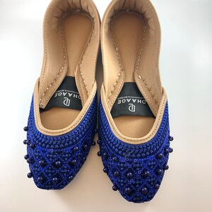 May include: A pair of blue embroidered flat shoes with beaded embellishments. The shoes have a tan leather interior and a black label with the text "DHAAGE London".