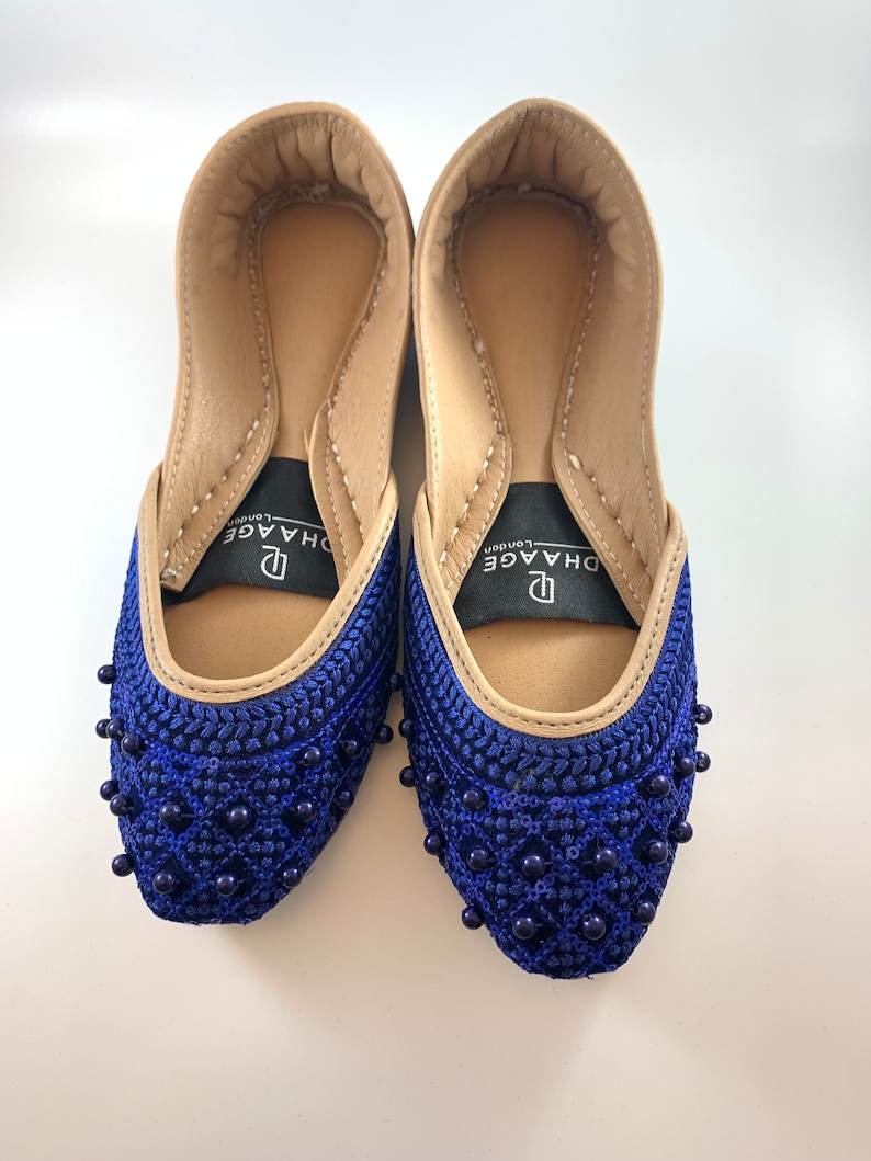 May include: A pair of blue embroidered flat shoes with beaded embellishments. The shoes have a tan leather interior and a black label with the text "DHAAGE London".