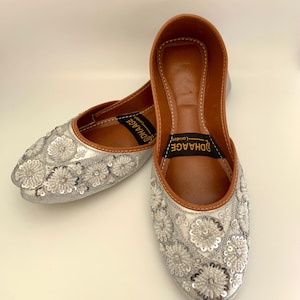 Peut inclure: Une paire de chaussures plates argent&eacute;es brod&eacute;es avec un int&eacute;rieur en cuir marron. Les chaussures ont une pointe pointue et sont d&eacute;cor&eacute;es de paillettes argent&eacute;es en forme de fleurs. Le nom de la marque "UDHAGE" est imprim&eacute; &agrave; l'int&eacute;rieur des chaussures.
