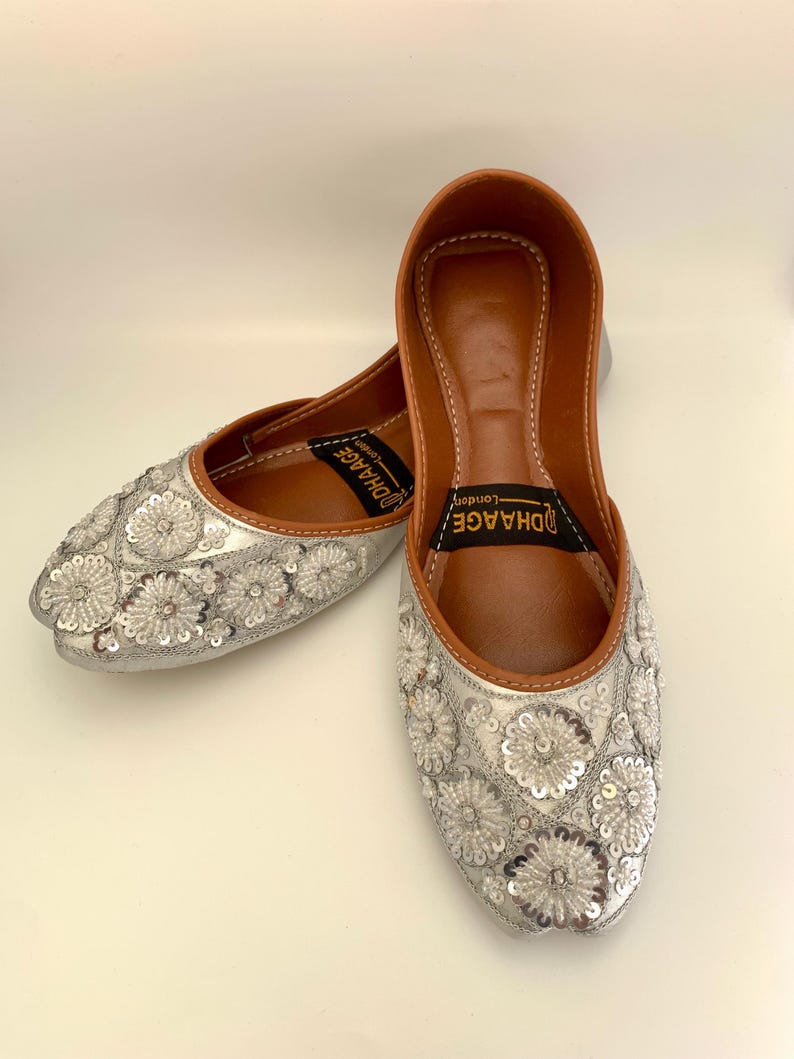 Peut inclure: Une paire de chaussures plates argent&eacute;es brod&eacute;es avec un int&eacute;rieur en cuir marron. Les chaussures ont une pointe pointue et sont d&eacute;cor&eacute;es de paillettes argent&eacute;es en forme de fleurs. Le nom de la marque "UDHAGE" est imprim&eacute; &agrave; l'int&eacute;rieur des chaussures.