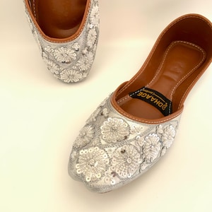 Peut inclure: Une paire de chaussures plates brod&eacute;es argent&eacute;es avec une semelle int&eacute;rieure en cuir marron. Les chaussures sont d&eacute;cor&eacute;es de sequins et de perles blancs en forme de fleurs. Le nom de la marque "DHAAGE" est imprim&eacute; sur la semelle int&eacute;rieure.