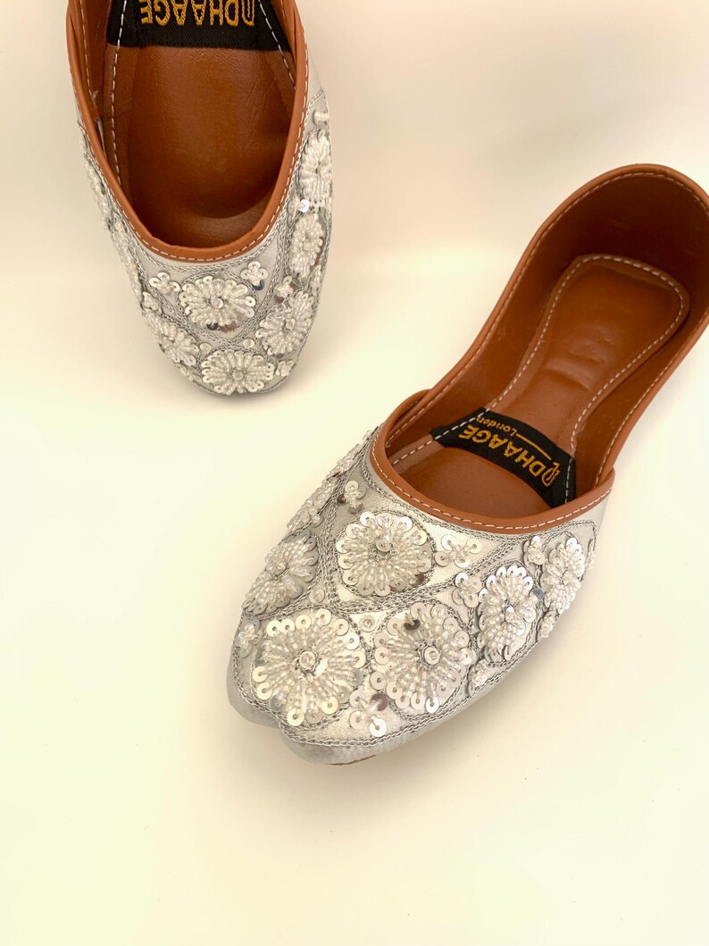 Peut inclure: Une paire de chaussures plates brod&eacute;es argent&eacute;es avec une semelle int&eacute;rieure en cuir marron. Les chaussures sont d&eacute;cor&eacute;es de sequins et de perles blancs en forme de fleurs. Le nom de la marque "DHAAGE" est imprim&eacute; sur la semelle int&eacute;rieure.