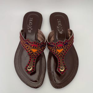 Könnte beinhalten: Ein Paar braune Ledersandalen mit bunter Stickerei um den Knöchelriemen. Die Sandalen haben eine spitze Zehenpartie und eine flache Sohle. Die Größe 10 ist auf der Innensohle jeder Sandale aufgedruckt.