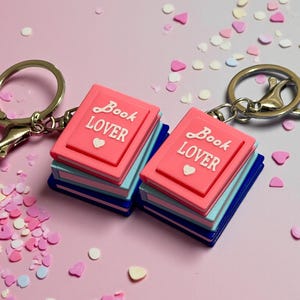 May include: Two book-shaped keyrings with pink covers and the words "Book Lover." The books have blue and light blue pages. The keyrings are silver and are on a pink background with confetti.