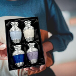 May include: A set of four decorative, small urns in a black box. Each urn has a silver-colored top and base with ornate detailing. The urns are in shades of blue, white, and purple. The box is held by a person wearing a blue and white striped sweater.