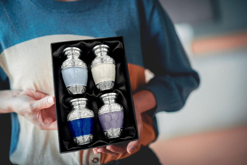 May include: A set of four decorative, small urns in a black box. Each urn has a silver-colored top and base with ornate detailing. The urns are in shades of blue, white, and purple. The box is held by a person wearing a blue and white striped sweater.