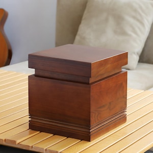 May include: A dark brown wooden square stool with a raised top and a decorative trim around the edges.