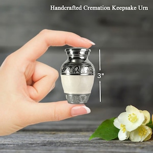 May include: Handcrafted cremation keepsake urn, 3 inches tall. The small urn is silver-toned with a white center section. The top and bottom have decorative patterns. The urn is held in a person's hand.