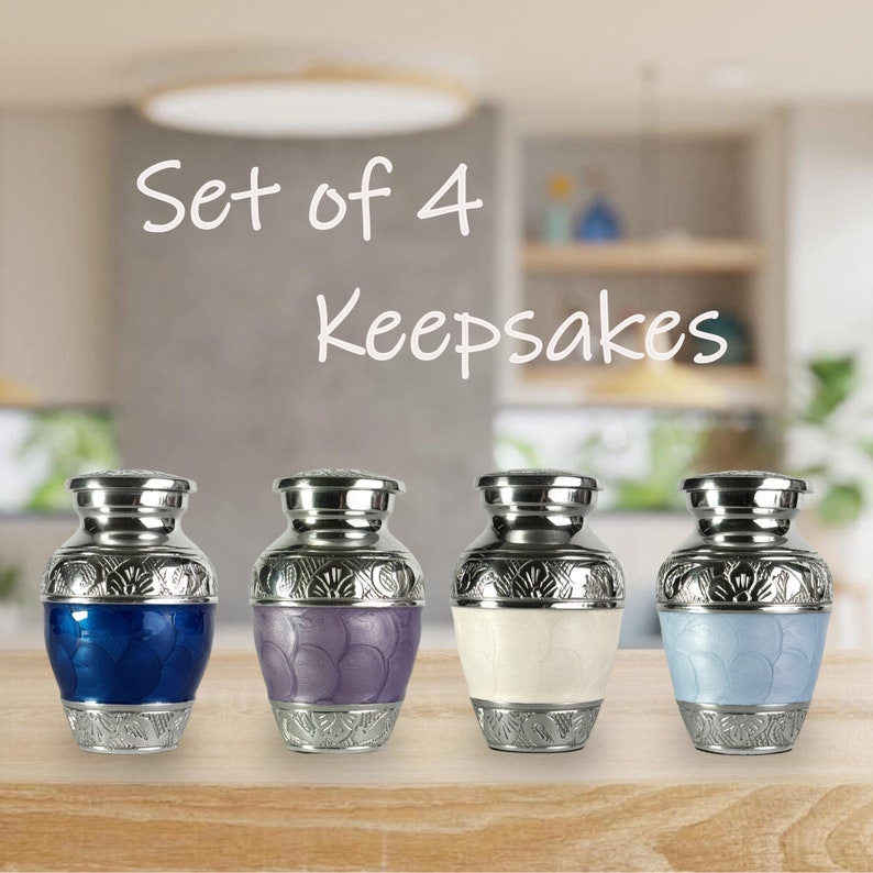 May include: Set of 4 keepsakes. Four small, decorative urns with silver-colored lids and bases. Each urn has a different colored enamel band: blue, purple, white, and light blue. The urns are displayed on a wooden surface.