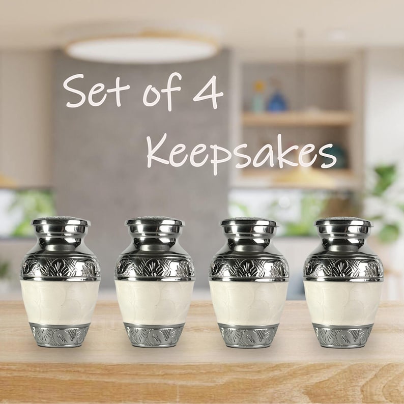 May include: Set of 4 keepsakes. Four small, decorative urns with silver-colored metal lids and bases. The urns have a cream-colored body with ornate silver-colored detailing. The text "Set of 4 Keepsakes" is in white script above the urns.