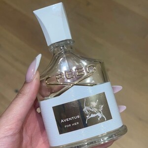 May include: A clear glass bottle of perfume with a white cap. The bottle has a gold label with the text "CREED" and "AVENTUS FOR HER".