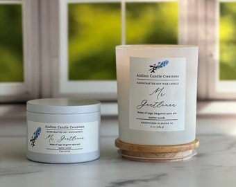 May include: Two scented soy wax candles, one in a silver tin and the other in a glass jar, both labeled "Mr. Gentleman." The labels include the maker's name and scent notes of sage, bergamot spice, and leather woods. The larger candle sits on a wooden coaster.