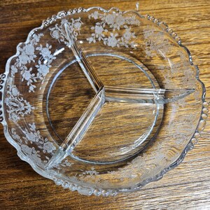 May include: A clear glass divided serving dish featuring three distinct sections separated by glass walls. The rim of the dish has a scalloped edge, and the surface is decorated with an etched floral pattern of roses and leaves around the perimeter.