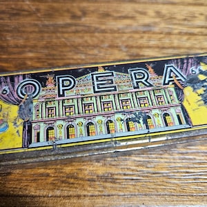 May include: A rectangular metal tin featuring a colorful illustration of an opera house building with the word OPERA printed in large black letters across the top. The design includes yellow, green, and purple accents with visible wear and scratches.