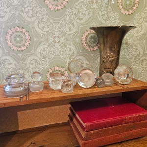 May include: A collection of clear glass bottle stoppers in various shapes and sizes arranged on a wooden shelf next to a tall, tarnished brass vase. The stoppers feature smooth, faceted, and rounded designs, with one small dark gray glass sphere.