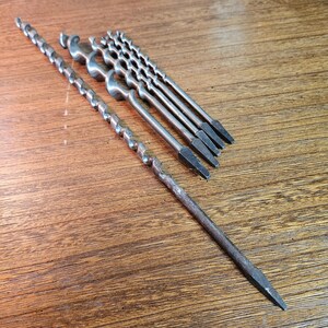 May include: A collection of six dark grey metal auger drill bits of varying lengths and diameters. Each bit features a spiral fluted shaft and a square tapered shank at the end, designed for manual drilling into wood with a brace or similar tool.